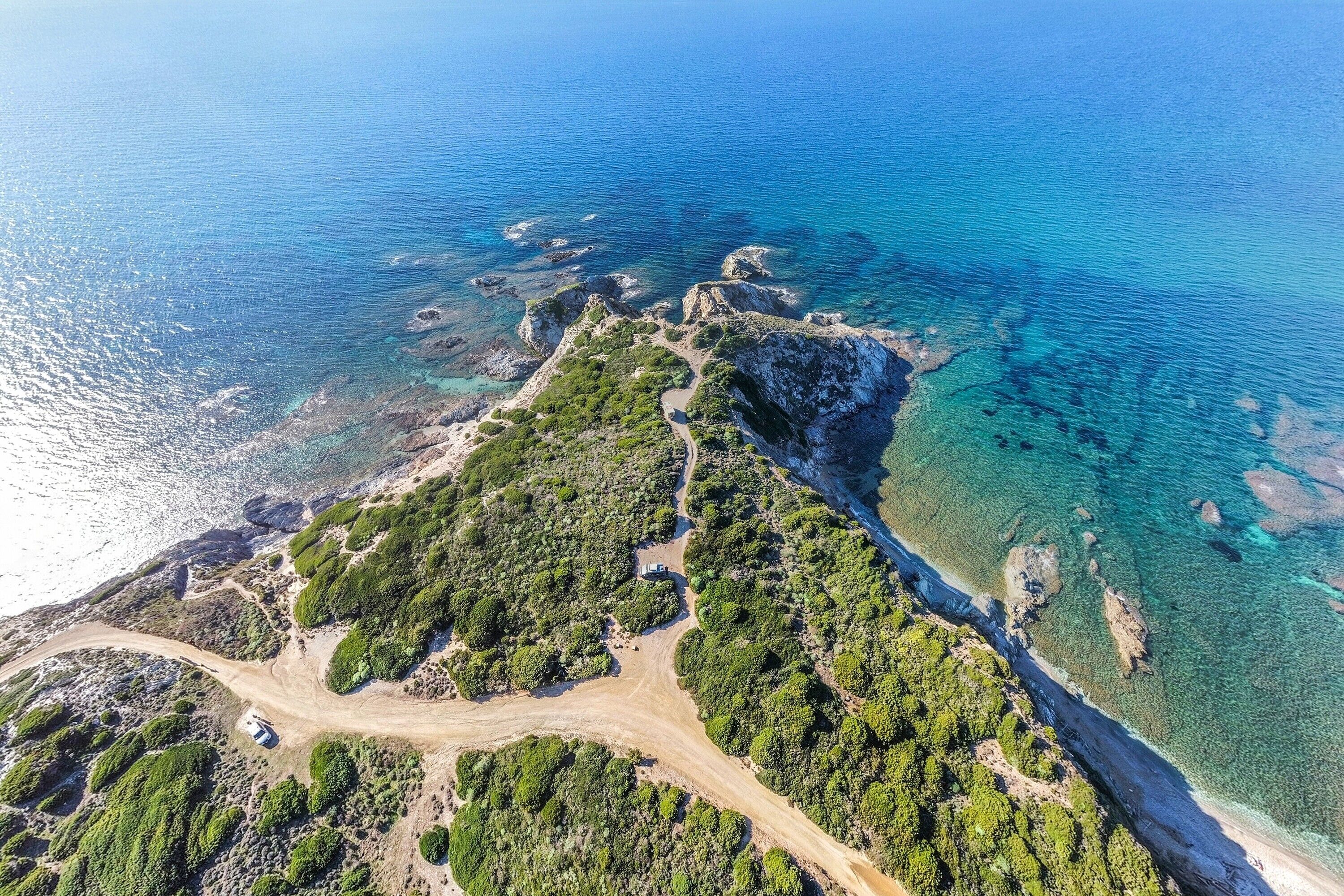 An aerial view of a coastal area with a dirt road, dense greenery, and clear blue waters.