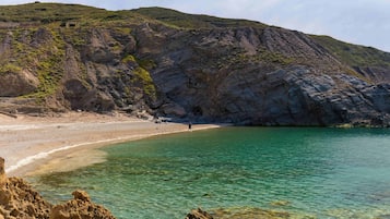A coastal landscape with a beach, clear water, rocky cliffs, and a person walking on the shore.