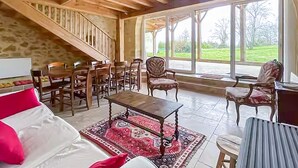 A spacious room with a wooden staircase, a dining area with chairs, a large window with a view of greenery, and a bed with a red pillow. - Belle maison avec piscine (Les Esseintes)