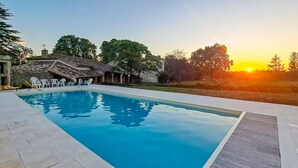 A swimming pool with a wooden deck, surrounded by trees and a building with a tiled roof. - Belle maison avec piscine (Les Esseintes)