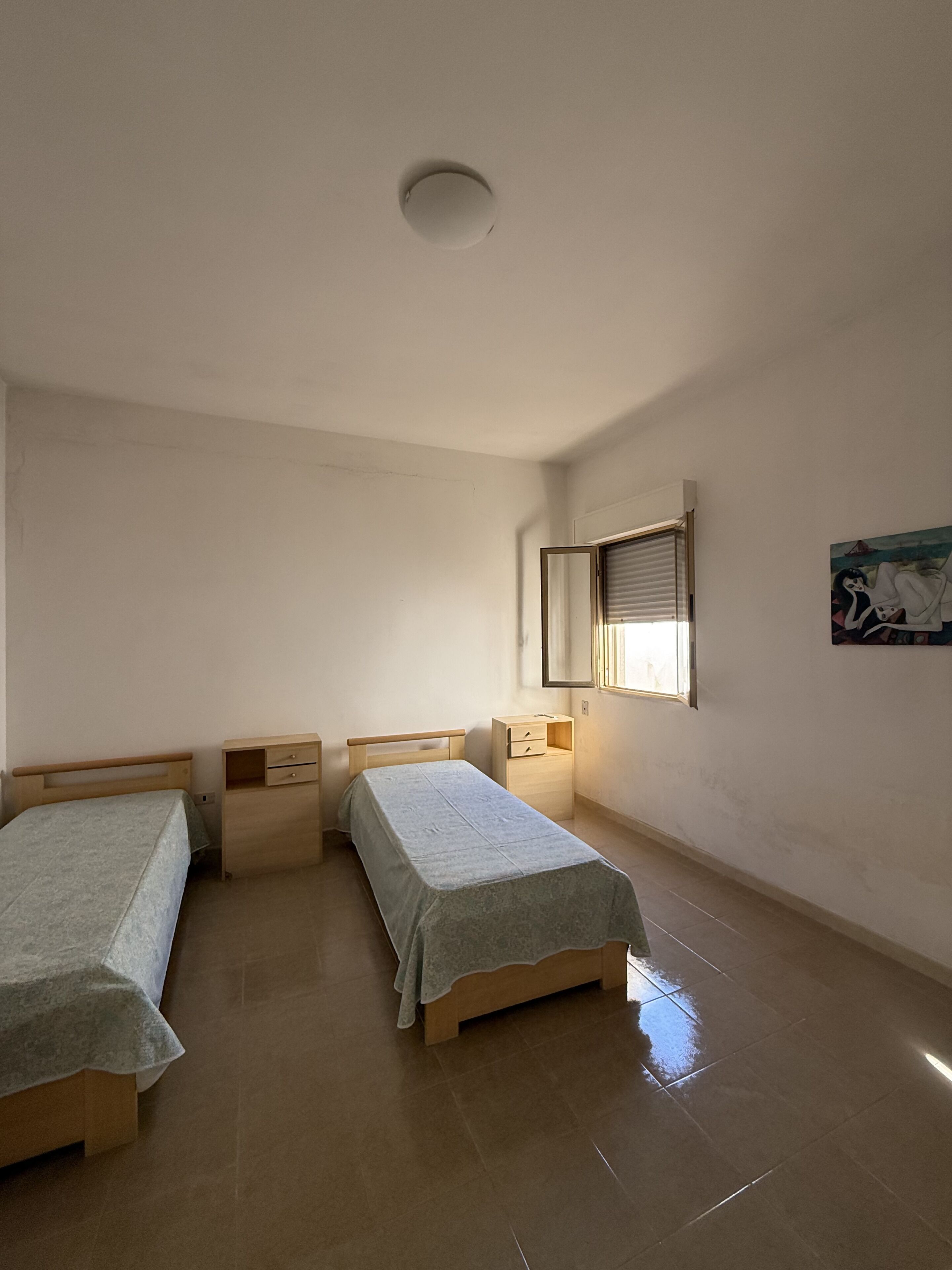 Une chambre avec deux lits simples, une table de chevet avec une lampe, une fenêtre avec des stores et un luminaire au plafond.