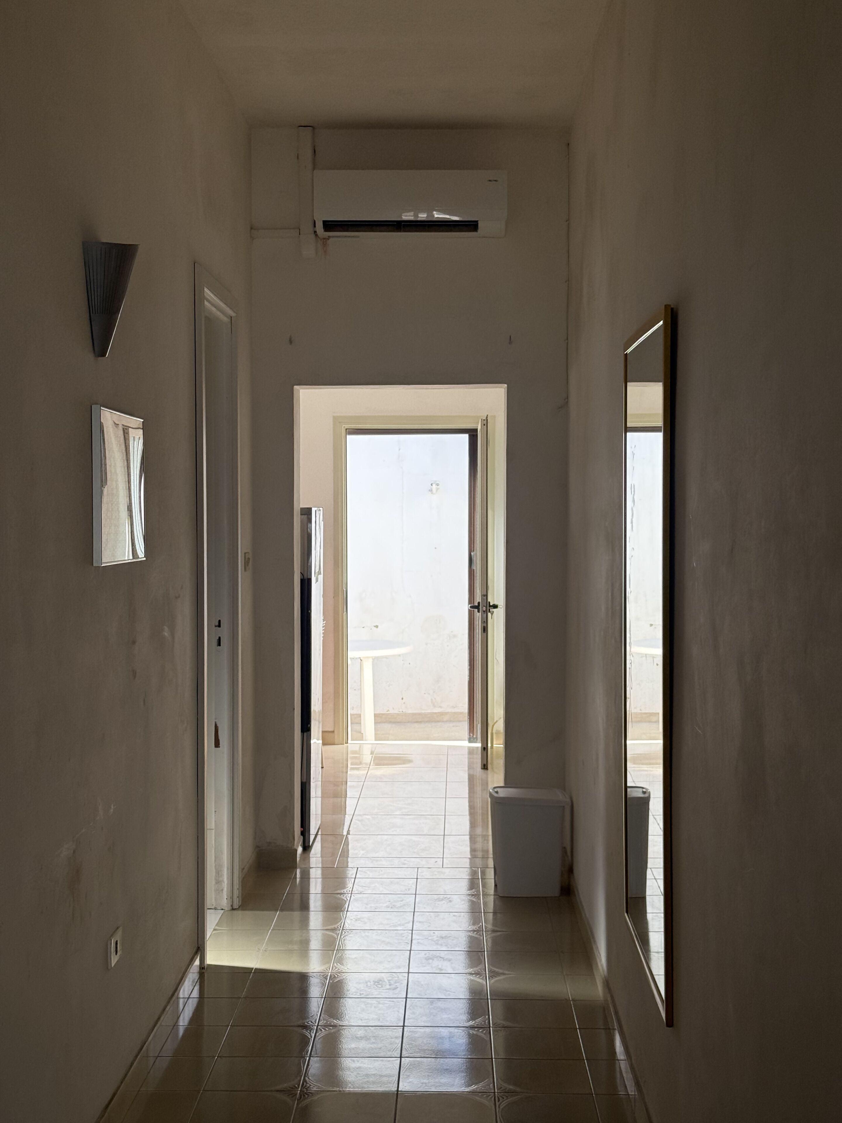 Um corredor com piso ladrilhado, uma porta que dá para uma varanda e um aparelho de ar condicionado na parede.