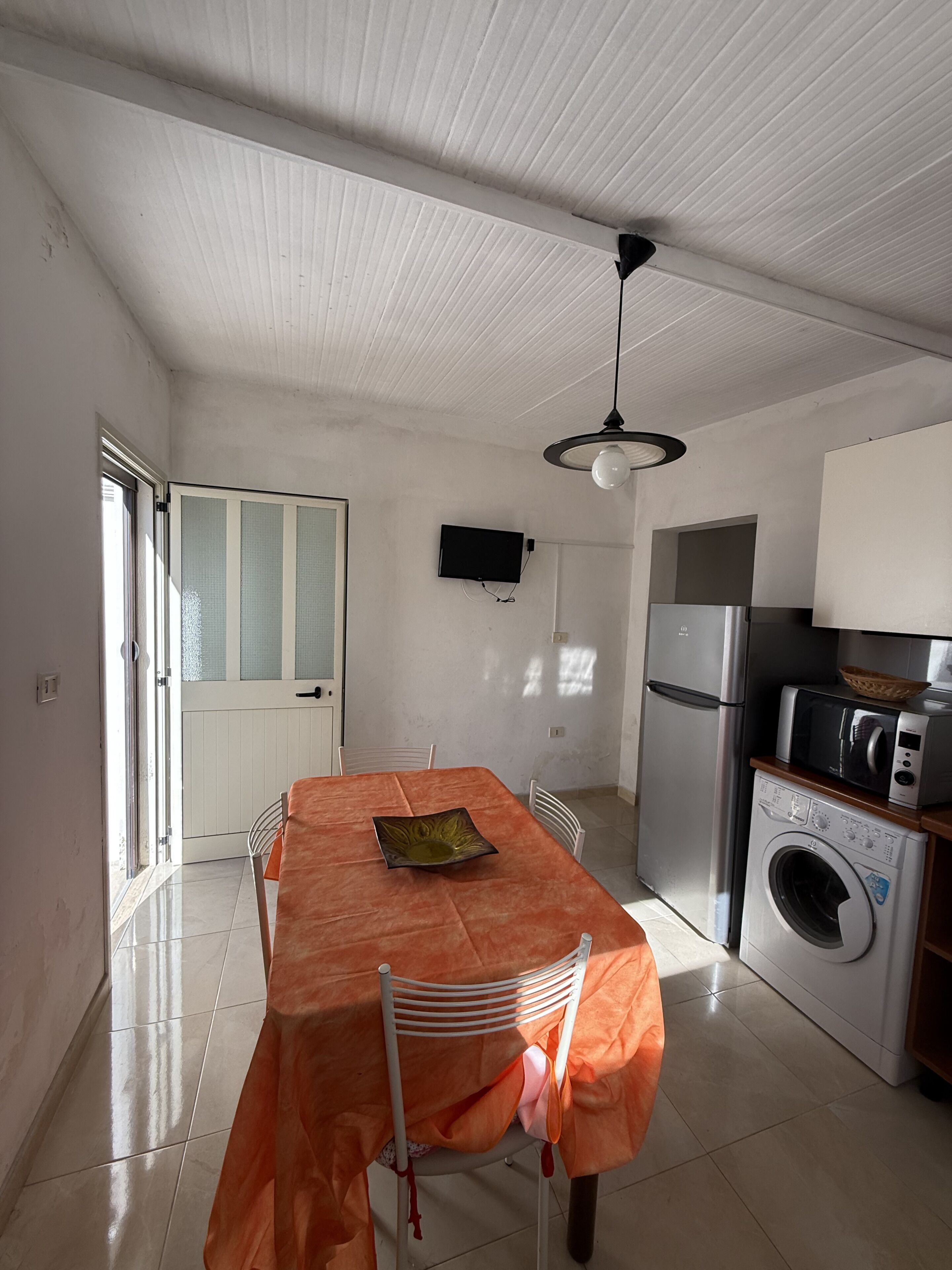 Una cocina con una mesa de comedor, sillas, lavadora, refrigerador y un televisor en la pared.