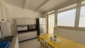 Una cucina compatta con mobili bianchi, un forno a microonde e un frigorifero. C'è un tavolo giallo con sedie e una finestra che lascia entrare luce naturale.