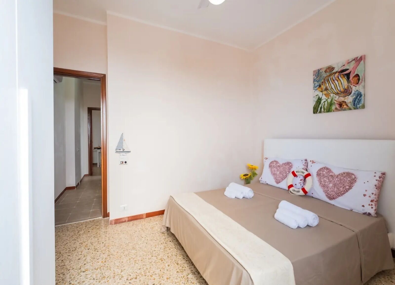 A bedroom with a bed, a painting, a heart-shaped pillow, and a small vase with flowers.