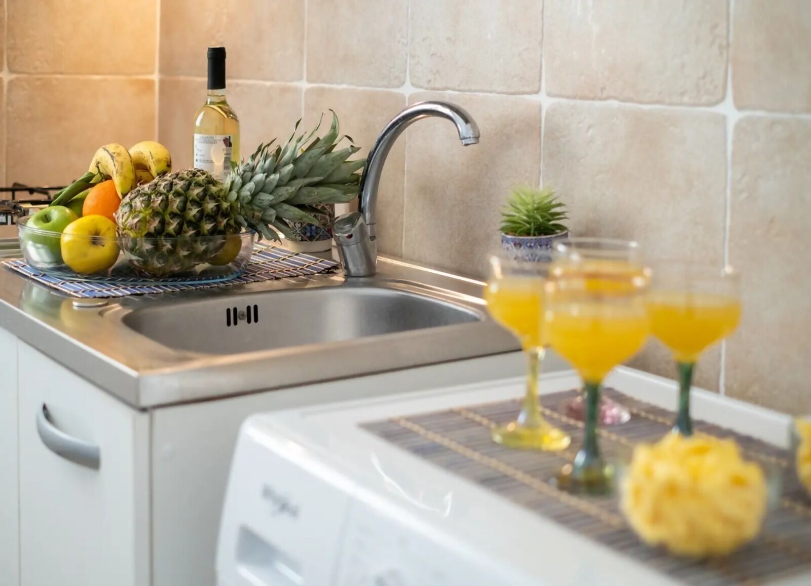 A kitchen counter with a sink, a bottle of white wine, a pineapple, and two glasses of orange juice.