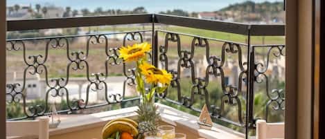Un balcón con una mesa dispuesta para el desayuno, que incluye frutas, un jarrón con girasoles y bebidas.