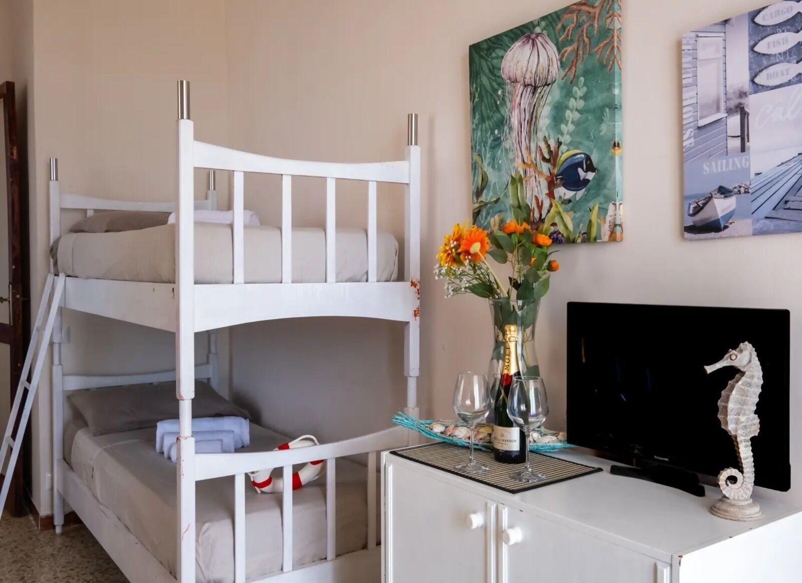 Una cama individual con escritorio, un florero con flores, una botella de vino y un adorno con forma de caballito de mar.