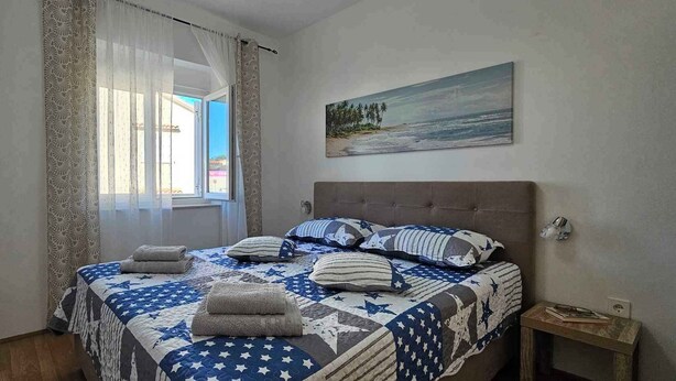 A bedroom with a bed, pillows, a nightstand, a painting, and a window with curtains. - Holiday flat with balcony (Šilo)
