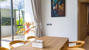 A dining area with a wooden table, chairs, a laptop, a tissue box, and a painting on the wall. - Aileen P3 V4 Saku Staycation (Sa Khu - Thalang)
