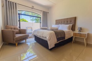 A modern bedroom with a bed, a chair, a bedside table, and a view of greenery outside.
