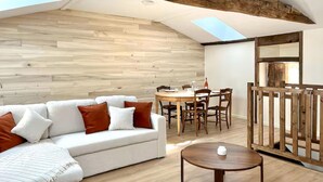 Ein Loftsesselzimmer mit einer Holzwand, einem weißen Sofa mit braunen Kissen, einem Esstisch mit Stühlen und einem kleinen Holztisch.