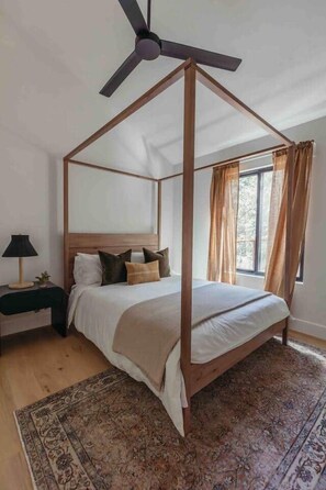 Una cama con dosel, un ventilador de techo, una mesita de noche con lámpara y una ventana con cortinas.