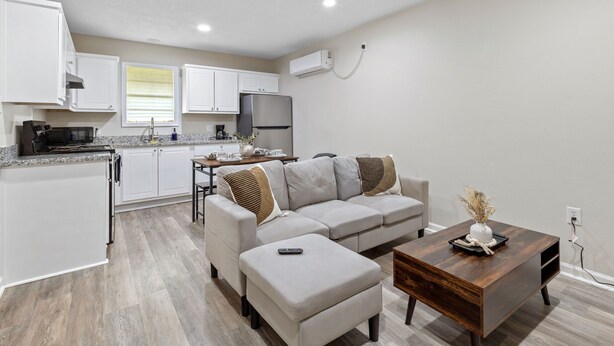 Una cocina moderna con armarios blancos, un refrigerador de acero inoxidable y una zona de comedor con sofá y reposapiés grises.