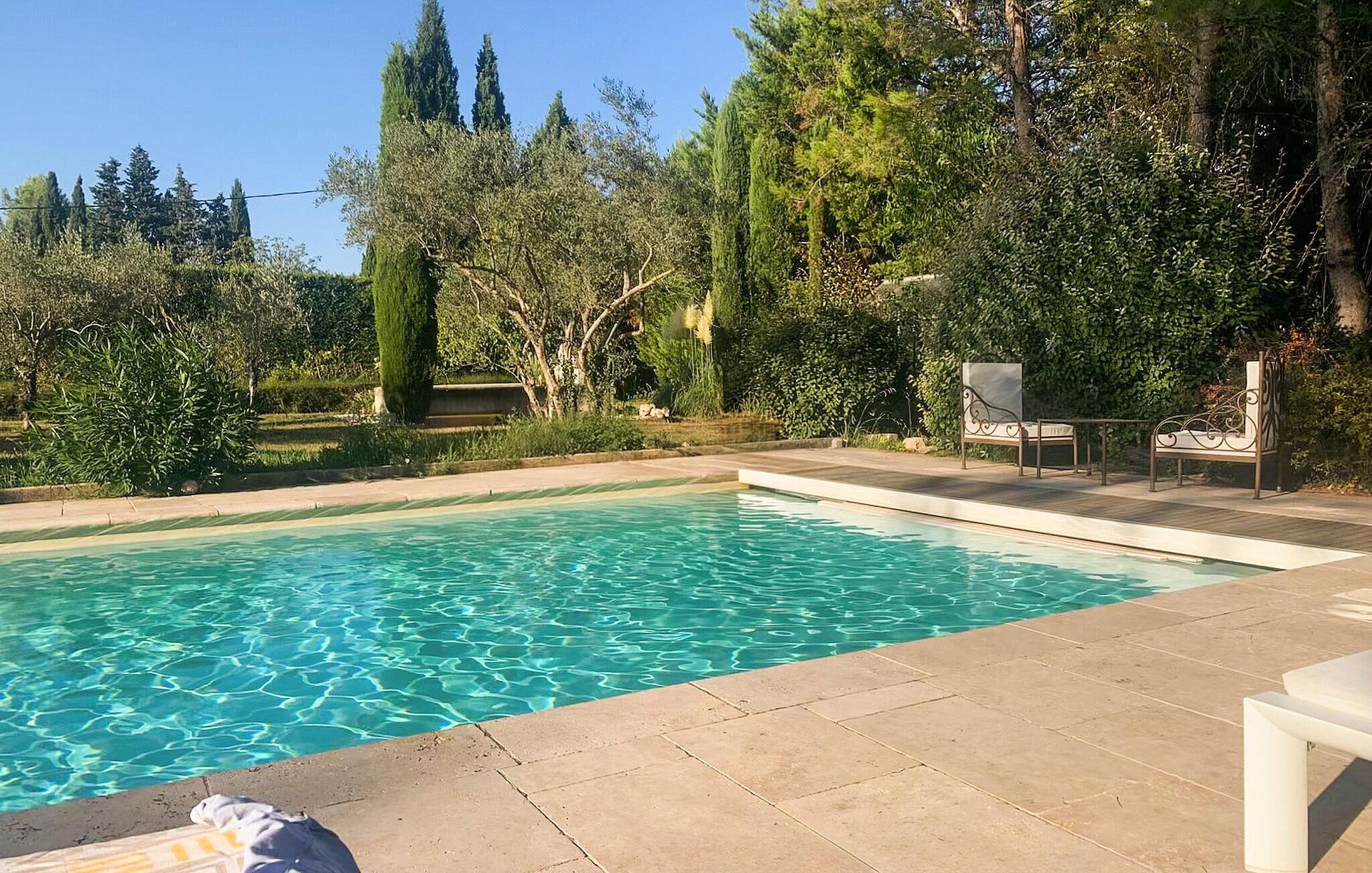A rectangular swimming pool with clear blue water, surrounded by a tiled deck and a variety of trees and shrubs.