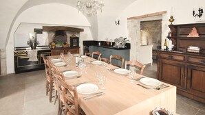 A dining area with a long table set for six, wooden chairs, a chandelier, a kitchen with a stone wall, and a cabinet with decorative items. - Maison avec piscine privée (Clansayes)
