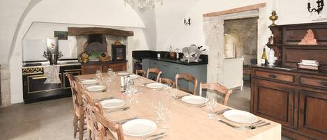 A dining area with a long table set for six, wooden chairs, a chandelier, a kitchen with a stone wall, and a cabinet with decorative items.
