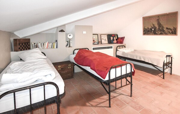 Une chambre de dortoir avec trois lits, un bureau et une étagère fixée au mur, où sont rangés des livres.
