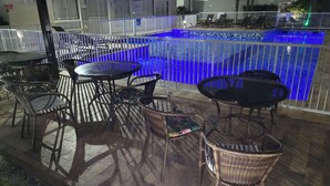 Un espace piscine avec une piscine illuminée de lumière bleue, un coin salon extérieur avec des tables et des chaises, et un périmètre délimité par une clôture.