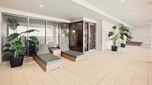 A spacious indoor area with a modern design, featuring a seating area with cushioned chairs and a sofa, large potted plants, and a glass partition. - Two Bedroom Unit in Central Adelaide (City of Adelaide)