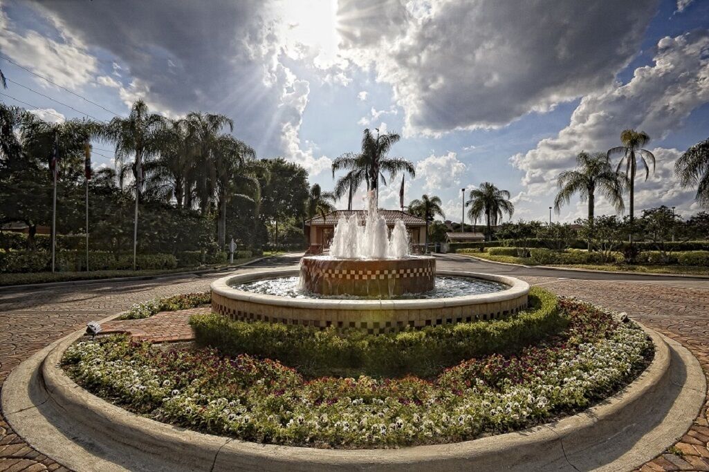 A circular fountain with water jets, surrounded by a landscaped garden with various plants and flowers, under a partly cloudy sky.