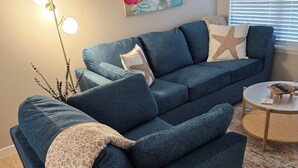 Een woonkamer met een blauwe bank, een vloerlamp, een schilderij en een raam met jaloezieën.