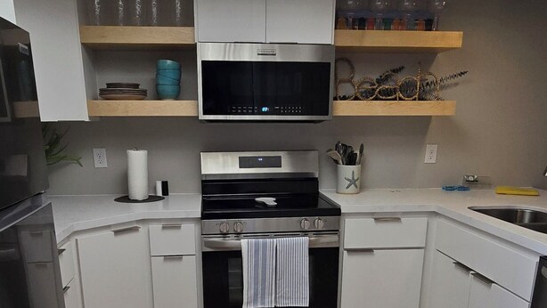 Een moderne keuken met witte kastjes, een inbouwoven, magnetron en vaatwasser. Boven de aanrecht zijn houten planken met verschillende spullen.
