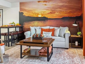 Un salon comprenant un canapé, une table basse et un grand tableau représentant un coucher de soleil sur l’eau.