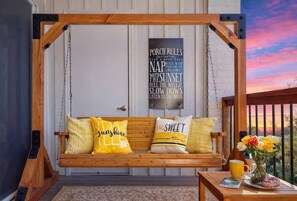 Un fauteuil suspendu en bois avec des coussins, une enseigne décorative pour le porche et une vue sur le coucher de soleil.