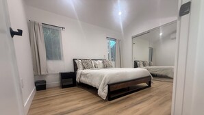 A bedroom with a wooden floor, a bed with a headboard, a nightstand, a mirror, and a window with curtains. - Perfect 2-bedroom corporate apartment with AC in peaceful Altadena (Altadena)