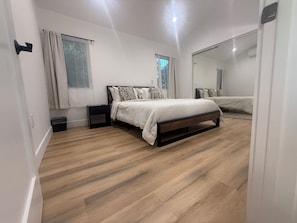 A bedroom with a wooden floor, a bed with a headboard, a nightstand, a mirror, and a window with curtains.