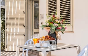 Ein Tisch mit einem Glas Orangensaft, einer Tasse und einem Tablett mit Gebäck, einer Vase mit Blumen sowie einem Fenster mit Rollläden.