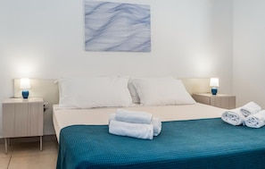 Ein Hotelzimmer mit einem Bett, zwei Nachttischen mit Lampen, einem Wandbild und einem ordentlich bezogenen Bett mit gefalteten Handtüchern.