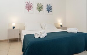 Ein Hotelzimmer mit einem Bett, weißen Leintüchern, einer türkisfarbenen Tagesdecke, zwei Nachttischlampen und drei korallenfarbenen Kunstwerken an der Wand.