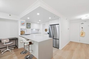 A modern kitchen with white cabinetry, a stainless steel refrigerator, and a bar area with white stools. - Victoria on Broadbeach (Broadbeach)