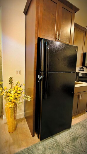 Une cuisine avec des meubles de rangement en bois, un réfrigérateur noir et un vase rempli de fleurs jaunes posé sur le sol.