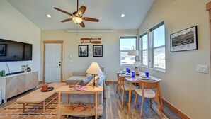 Sala de estar bem iluminada com ventilador de teto, área de jantar com mesa e cadeiras, e cozinha com televisão e balcão.