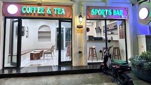 Une devanture avec les panneaux « Café et thé » et « Bar sportif », une moto garée devant, et un pot de fleurs sur le côté droit.