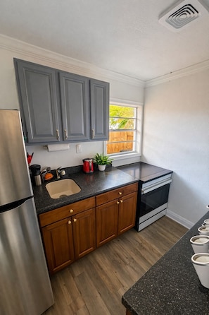 A kitchen with grey cabinets, a stainless steel refrigerator, a black countertop, a window with a view of a wooden fence, and a built-in oven. - Entire rental unit Studio in Altamonte Spring (Altamonte Springs)