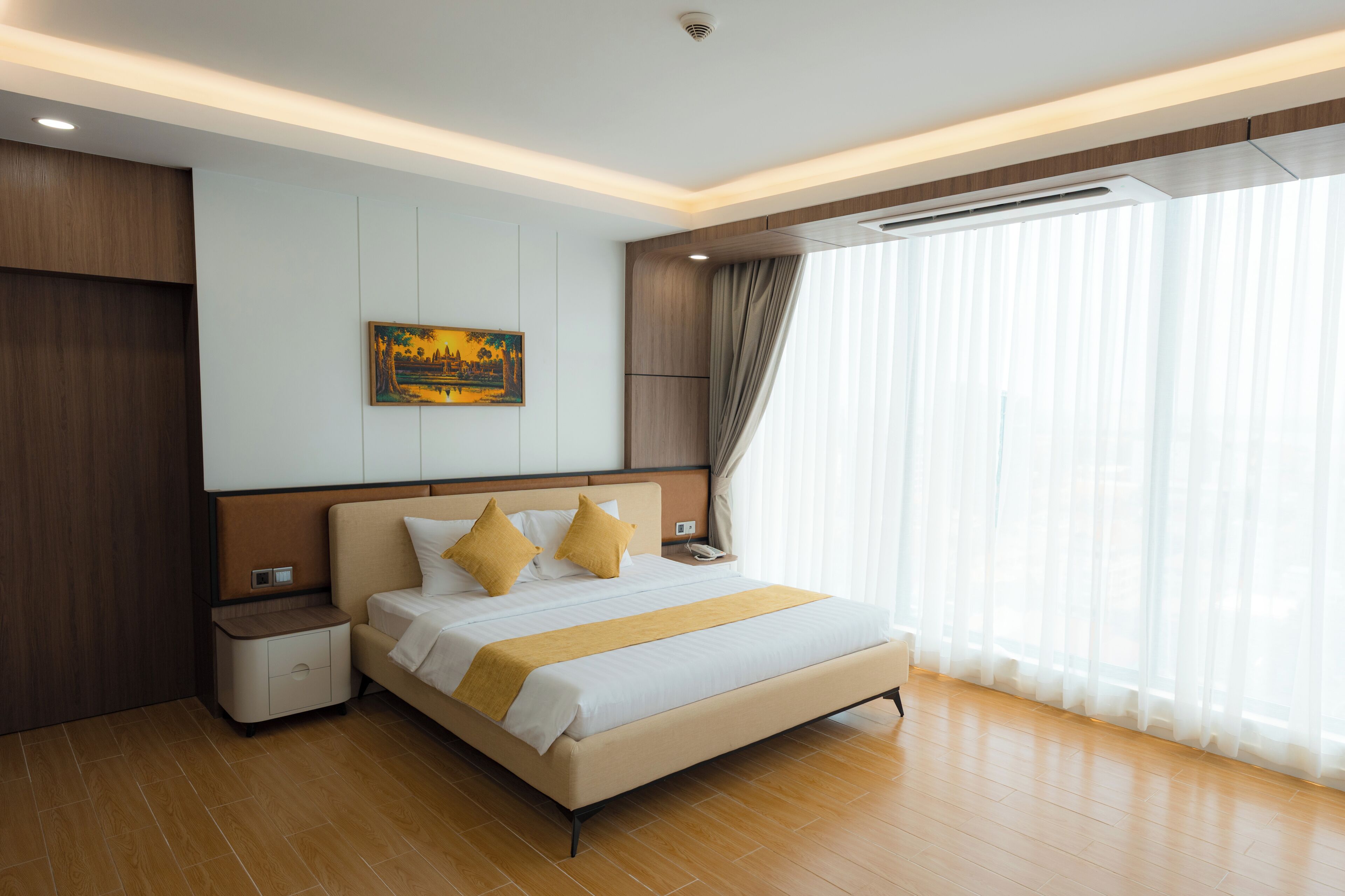 Een moderne hotelkamer met een groot bed, houten lambrisering, een schilderij aan de muur en een groot raam met dunne gordijnen.