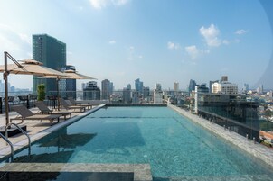 Piscina infinita en la azotea con sillones de descanso y sombrillas, con vistas a un horizonte urbano.