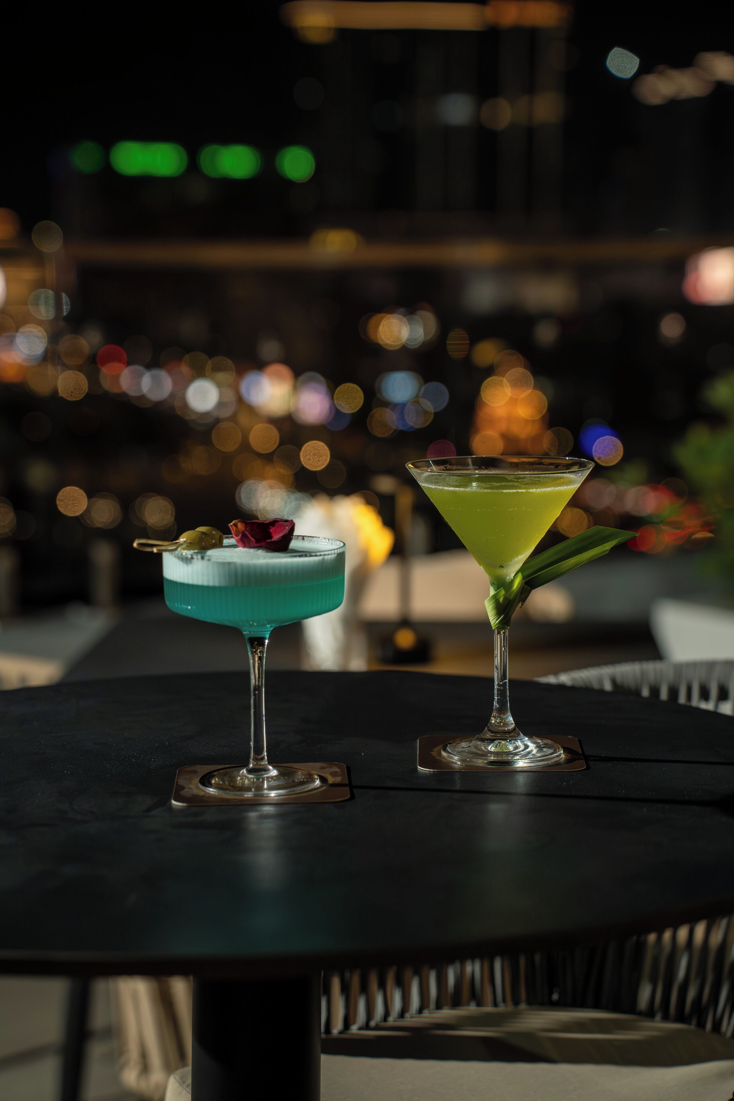 Two cocktails on a table with a blurred cityscape in the background.