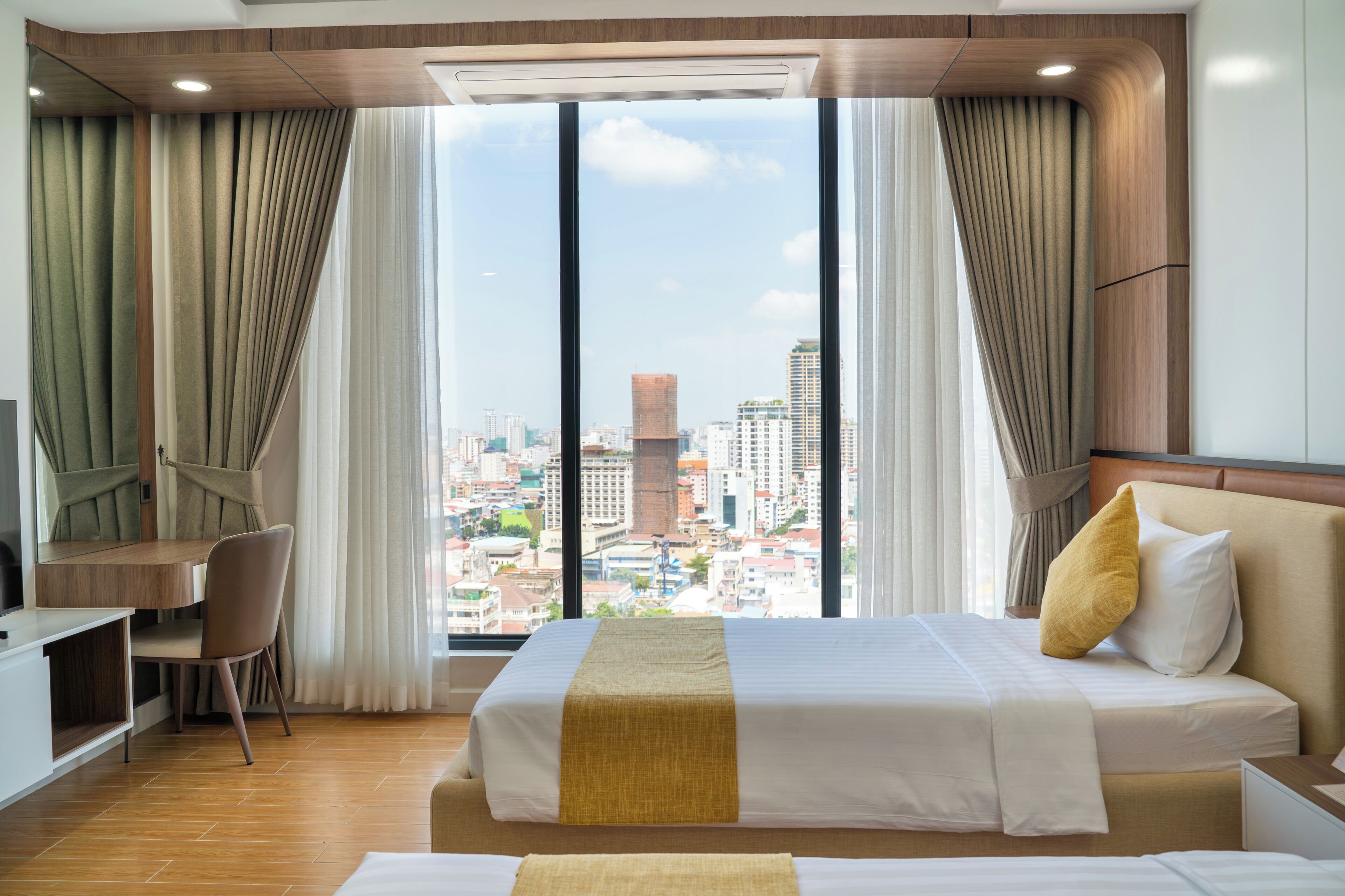 A hotel room with a bed, a desk, and a chair. There is a large window with curtains, and a city view is visible.