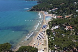 An aerial view of a coastal area with a beach lined with rows of umbrellas and beach chairs, indicating a resort or hotel area. - CASA MANIDU - TRILOCALE LAVANDA (Capoliveri)