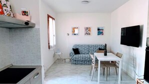 Een compacte keuken met een aanrecht en kastjes, een eethoek met tafel en stoelen, een bank en een televisie die aan de muur hangt.