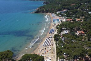Eine Luftaufnahme einer Küstenlandschaft mit einem Strand, der mit Reihen von Sonnenschirmen und Liegestühlen gesäumt ist, was auf ein Resort oder Hotel hindeutet.