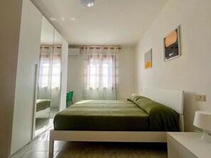 A bedroom with a bed, a chair, a bedside table, and a window with curtains. - CASA MANIDU - TRILOCALE OLIVA (Capoliveri)