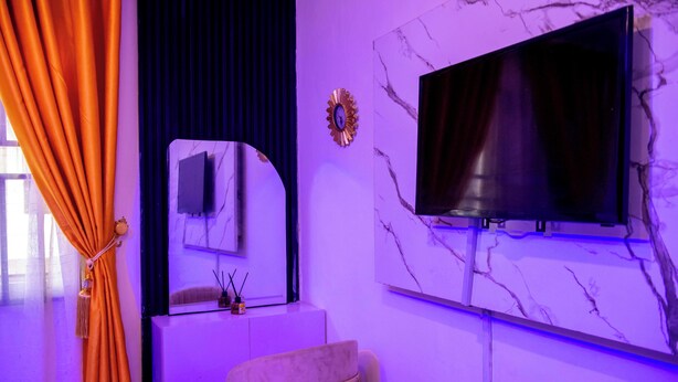 A room with a TV mounted on a marble wall, a mirror, and a window with curtains. - J3's Apartment & Lounge (Benin City)