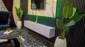 A modern living room with a TV mounted on a marble wall, a black sofa, a white console table, and large green plants. - J3's Apartment & Lounge (Benin City)