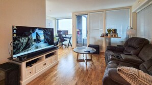 Sala de estar com televisão de tela plana, sofá, mesa de centro e porta de vidro deslizante que dá para uma varanda.